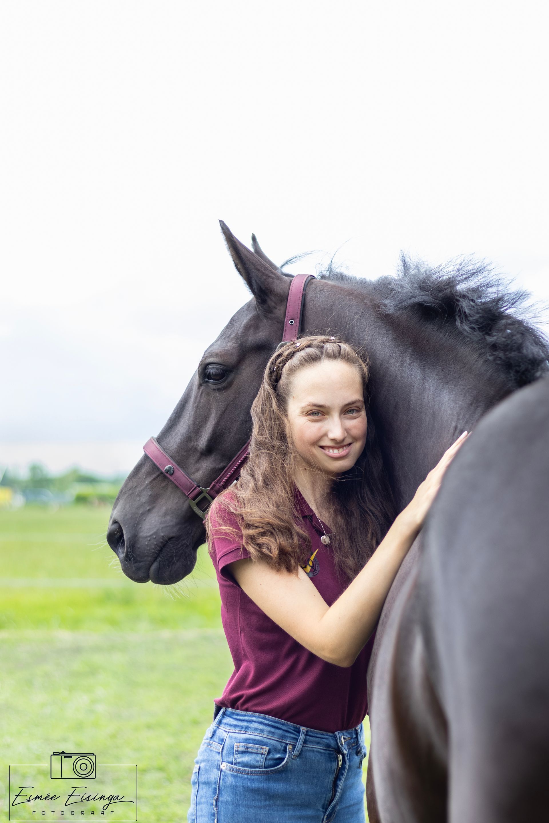 Vrouw glimlacht en omhelst een zwart paard in een veld. Ze draagt een bordeauxrood shirt en een spijkerbroek.