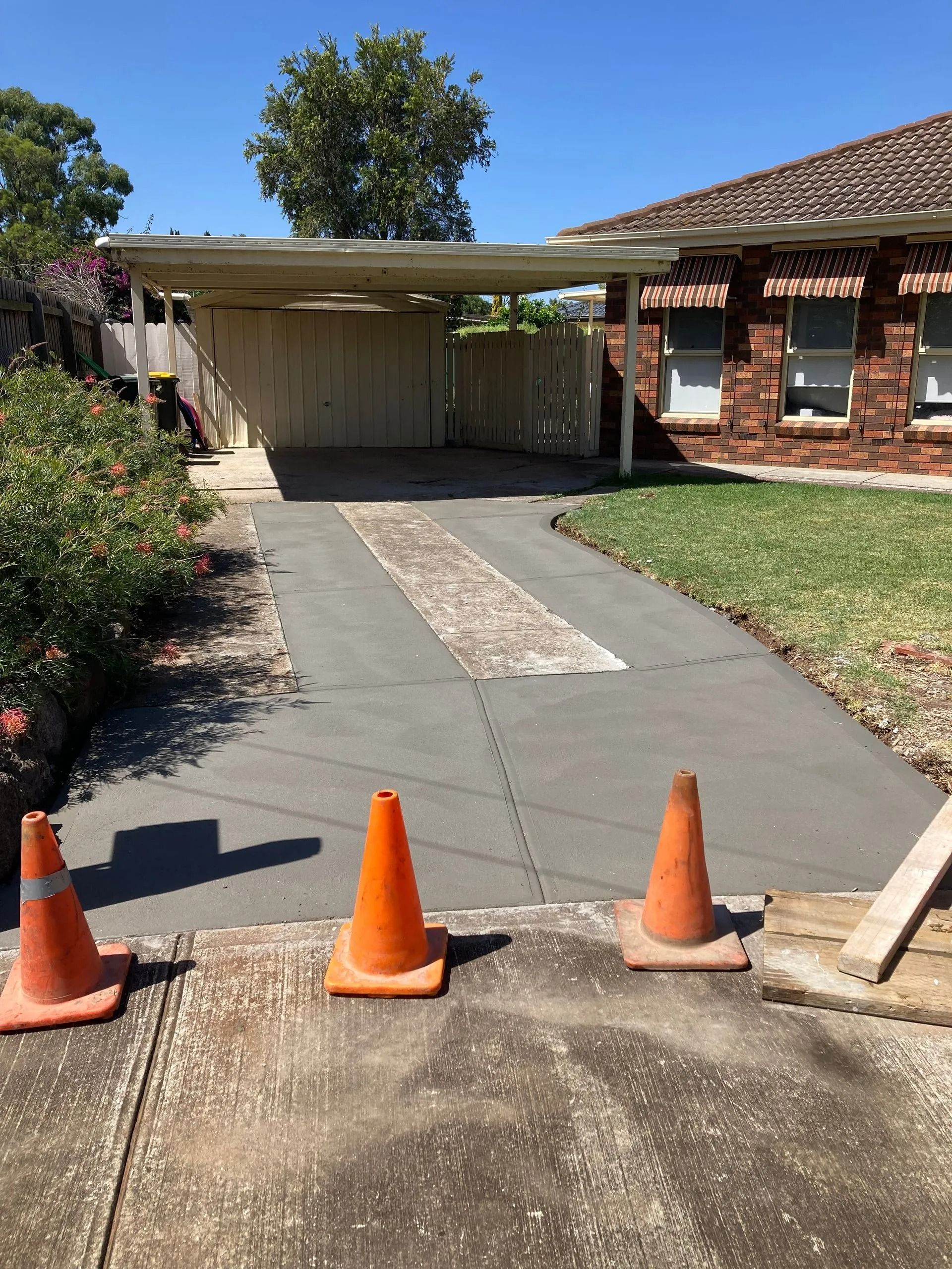 Driveway under construction, with orange cones. Cement, carport, brick house, and greenery are visible. Sunny day.