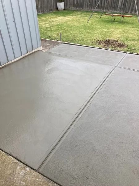 Newly poured gray concrete patio with control joints, next to a corrugated metal wall and a grassy yard.
