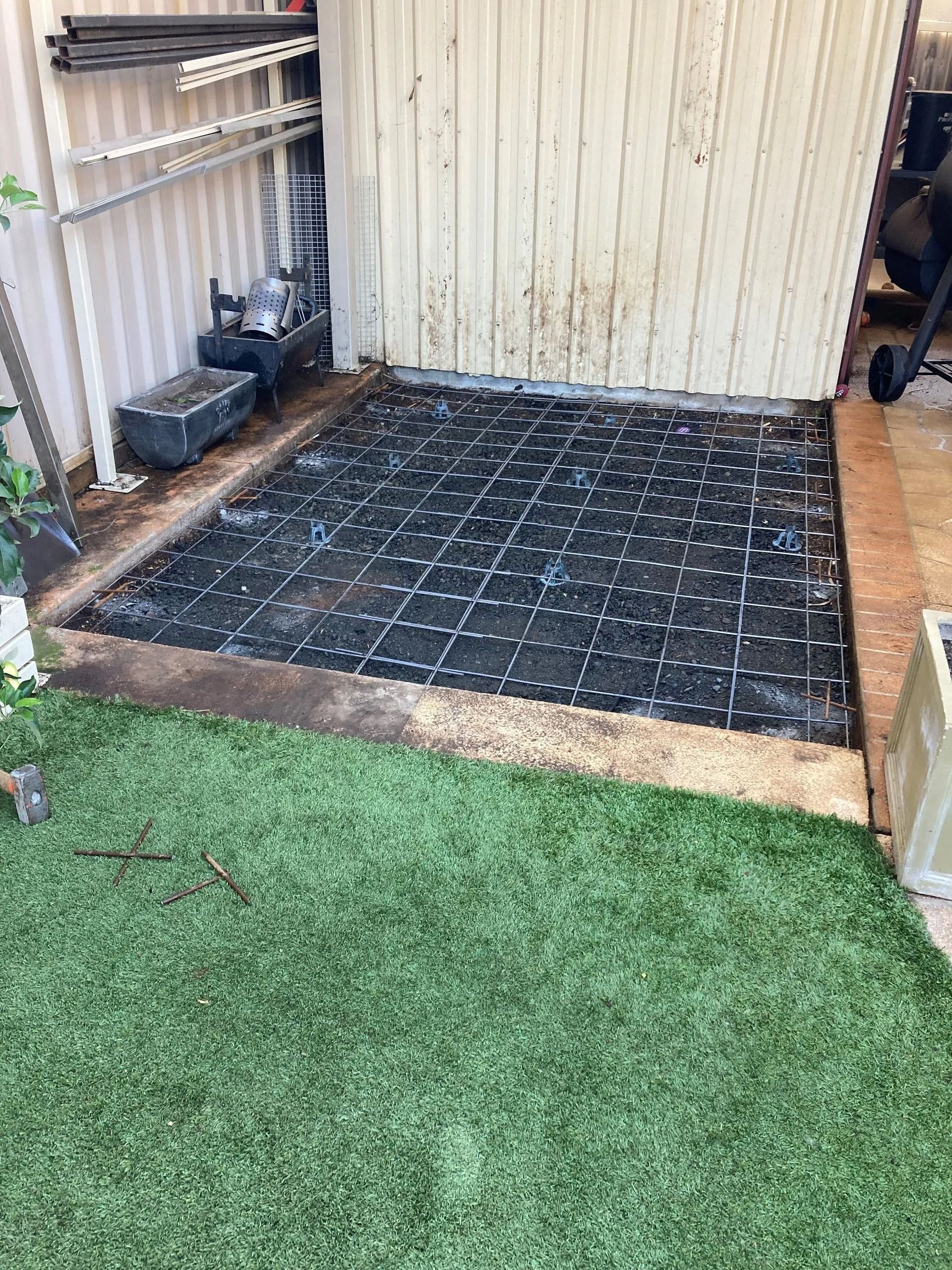 Black grid on a concrete base, bordered by wood and grass, next to a shed wall.
