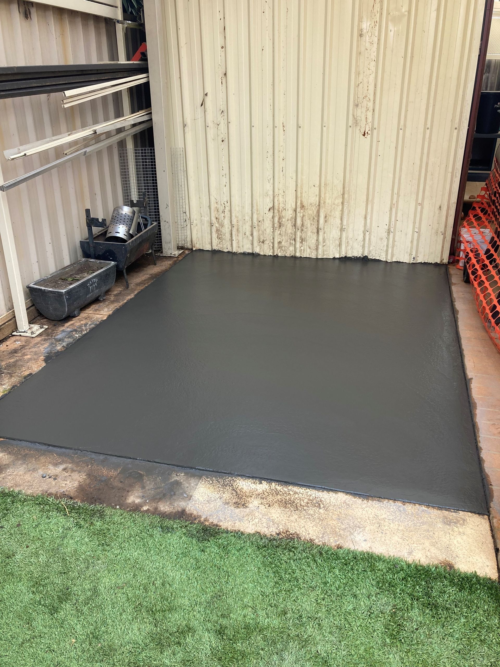 Newly poured dark gray concrete slab next to a light-colored wall and artificial grass.
