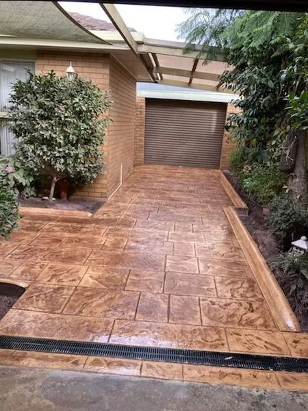 Stamped concrete driveway leading to a garage. Brown with tile pattern, surrounded by landscaping.