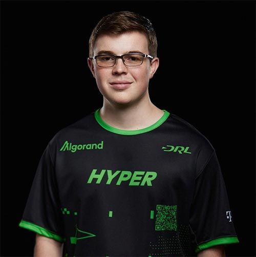 A young man wearing glasses and a hyper shirt is standing in front of a black background.