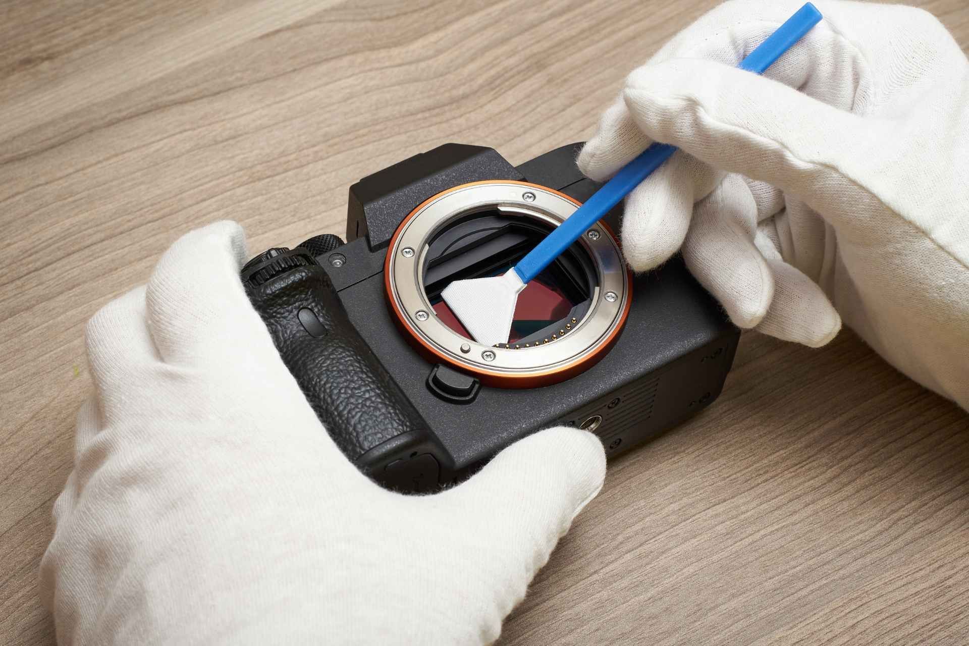 Sensor Cleaning image, image of hand holding camera lens, brushing in with a soft brush to clean it, with jar of qtips out of focus on a wooden table in the background