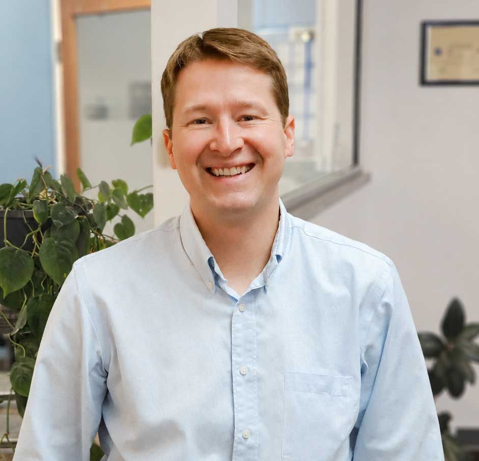 Smiling person in a light blue shirt standing in an office hallway with plants in the background Smiling person in a light blue shirt standing in an office hallway with plants in the background