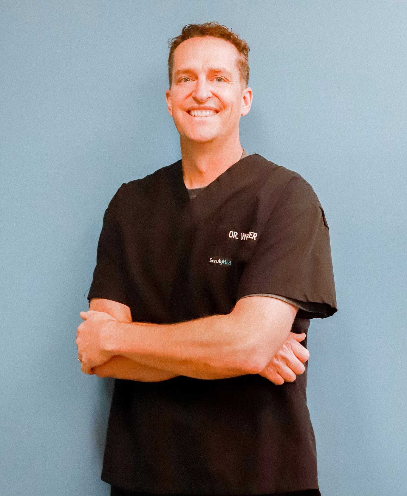 Smiling person in a black scrub top with arms crossed against a blue wall Smiling person in a black scrub top with arms crossed against a blue wall
