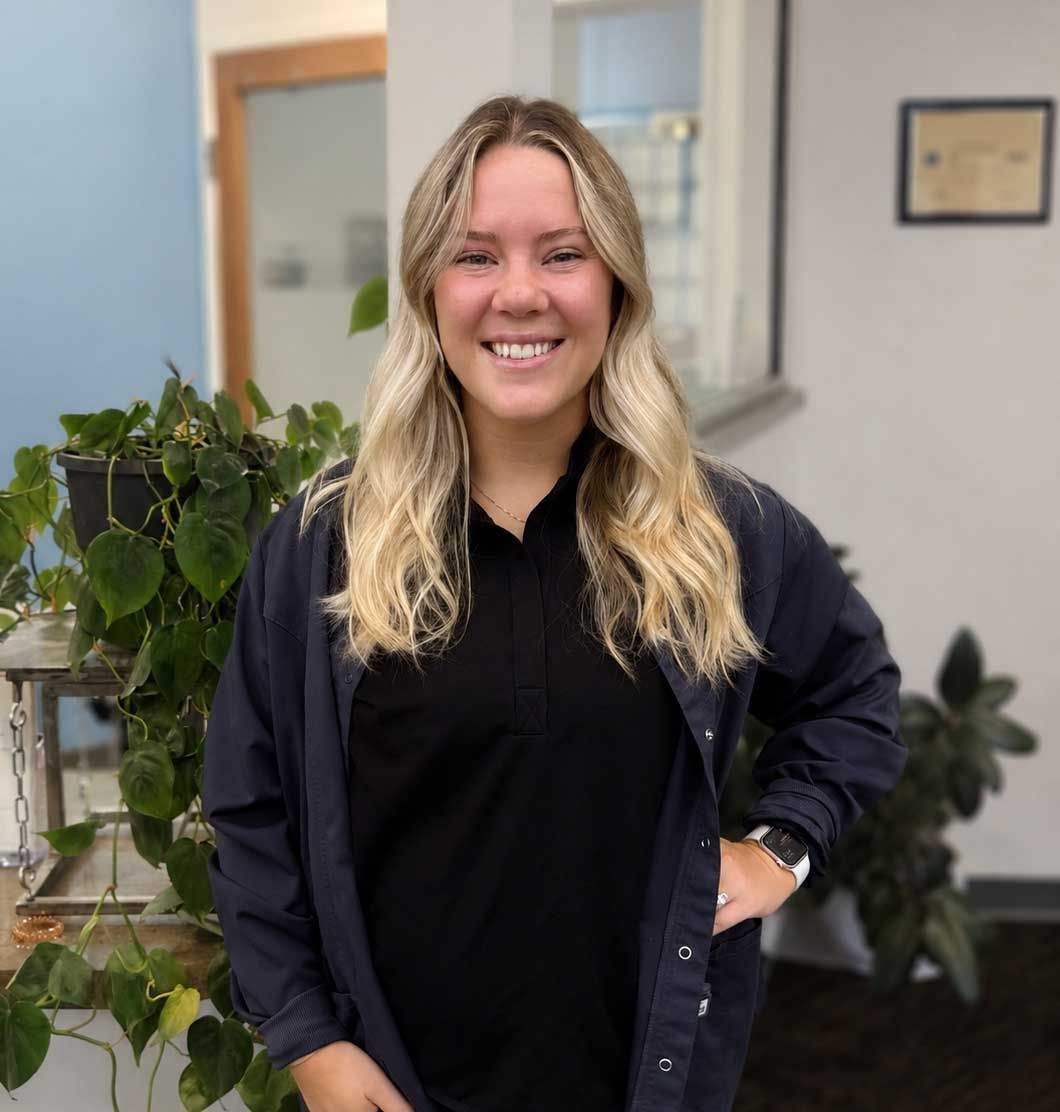 Smiling person in a black shirt and navy jacket stands indoors near plants and office windows. Smiling person in a black shirt and navy jacket stands indoors near plants and office windows.