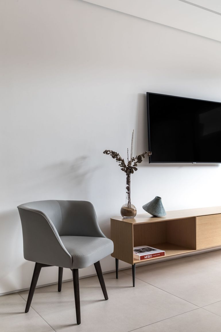 Uma poltrona cinza fica ao lado de um móvel de madeira clara para aparelhos eletrônicos, sob uma TV fixada na parede, em uma sala minimalista com paredes brancas.