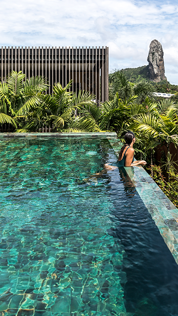 Uma pessoa contempla uma formação rochosa imponente enquanto descansa em uma piscina exuberante de borda infinita em um resort.
