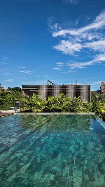 Uma piscina infinita de águas cristalinas reflete um céu azul brilhante, situada em frente a um edifício moderno rodeado por uma vegetação exuberante.