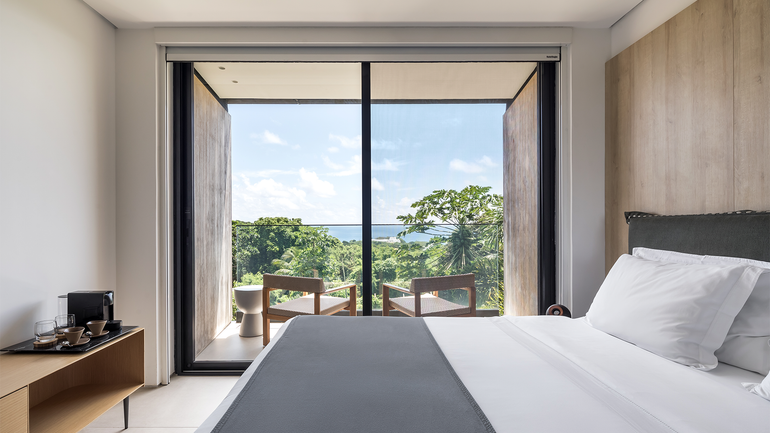 Um quarto de hotel luminoso com uma cama de lençóis brancos e uma grande janela com vista para as árvores e o oceano ao longe.