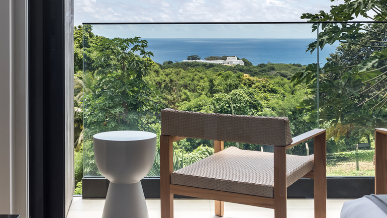 Uma cadeira de madeira para área externa e uma mesa lateral branca em uma varanda com vista para uma paisagem verdejante e o oceano.