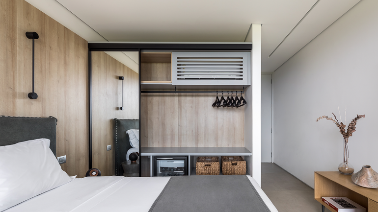 Um quarto de hotel moderno com cama, guarda-roupa aberto com prateleiras, cofre e cabides, ao lado de uma escrivaninha minimalista de madeira.