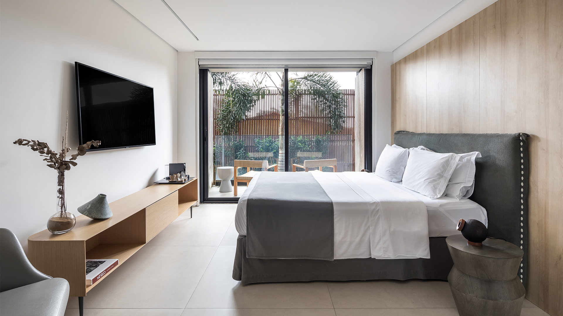 Una habitación de hotel moderna con cama de matrimonio, pared revestida de madera, televisor sobre una consola de madera clara y puerta de cristal que da a un patio privado.