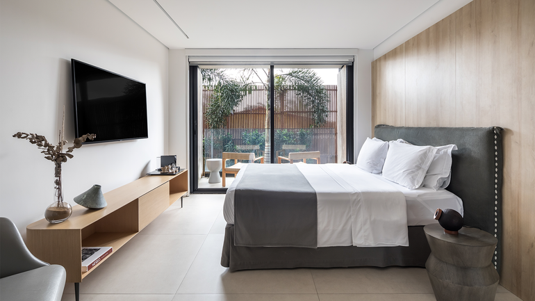 Um quarto de hotel moderno e minimalista com cama, móvel de madeira para equipamentos de mídia, TV de parede e varanda com área de estar externa.