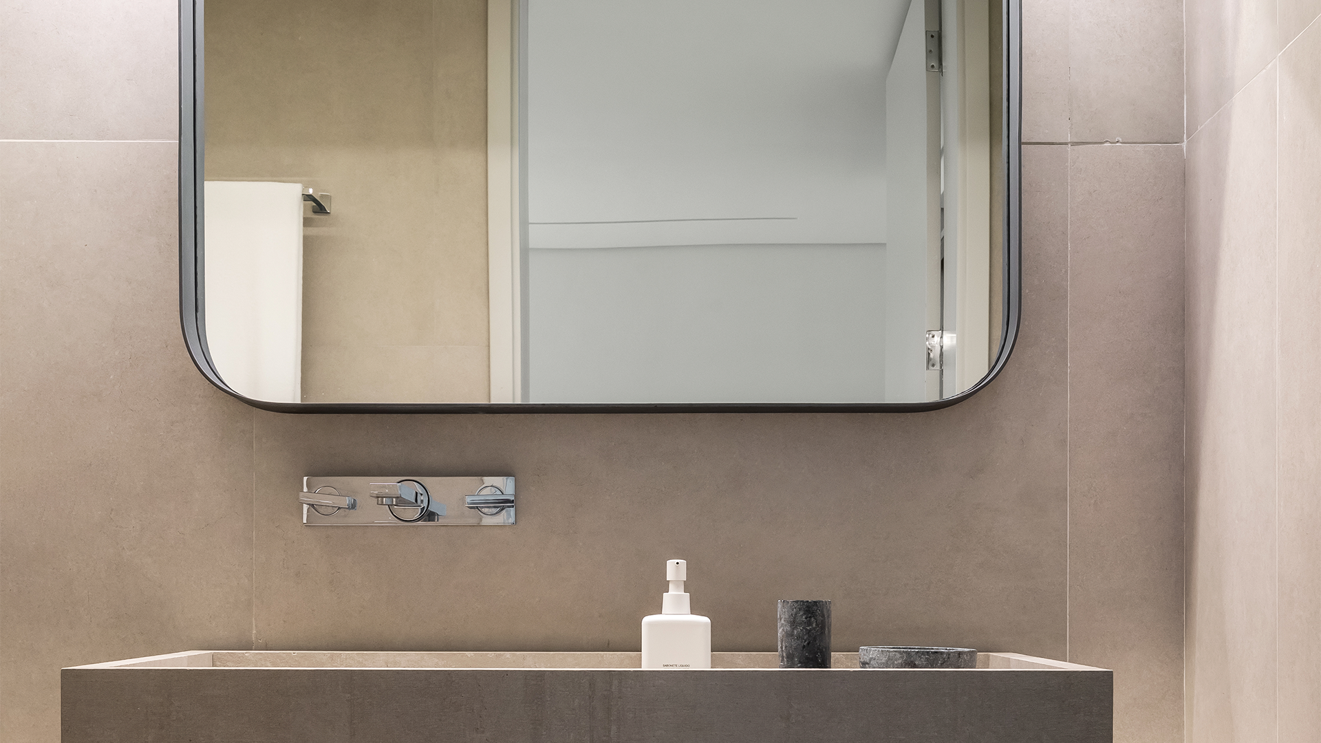 Tocador de baño moderno con lavabo de piedra gris, grifo cromado montado en la pared, espejo rectangular y dispensadores de jabón.