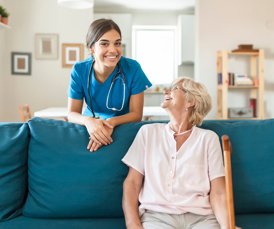 A smiling caregiver in blue scrubs leaning over a couch toward a seated senior with a cane in a home setting.
