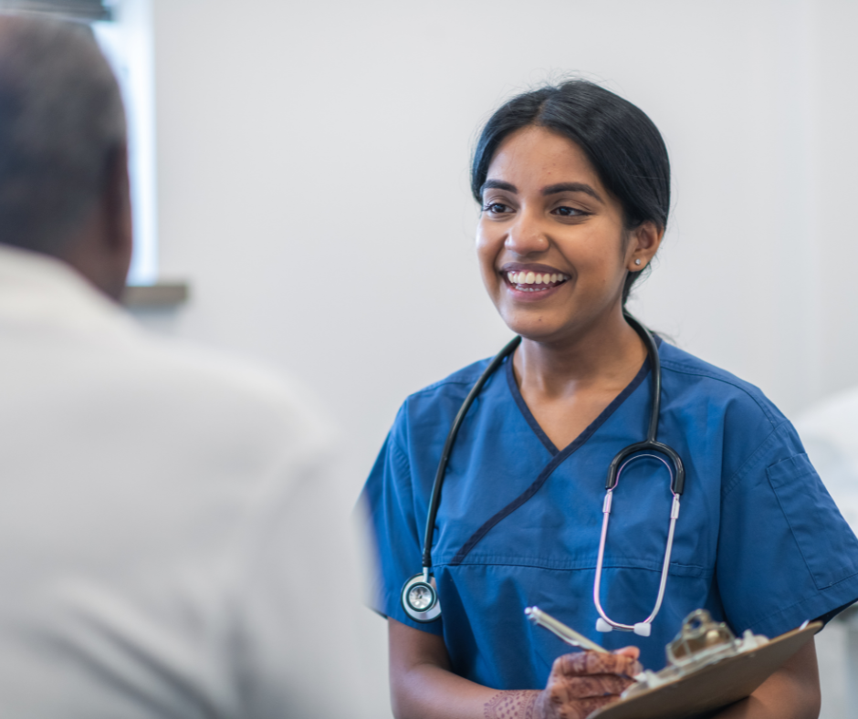 A healthcare professional in blue scrubs with a stethoscope smiles while holding a clipboard, speaking to a patient.