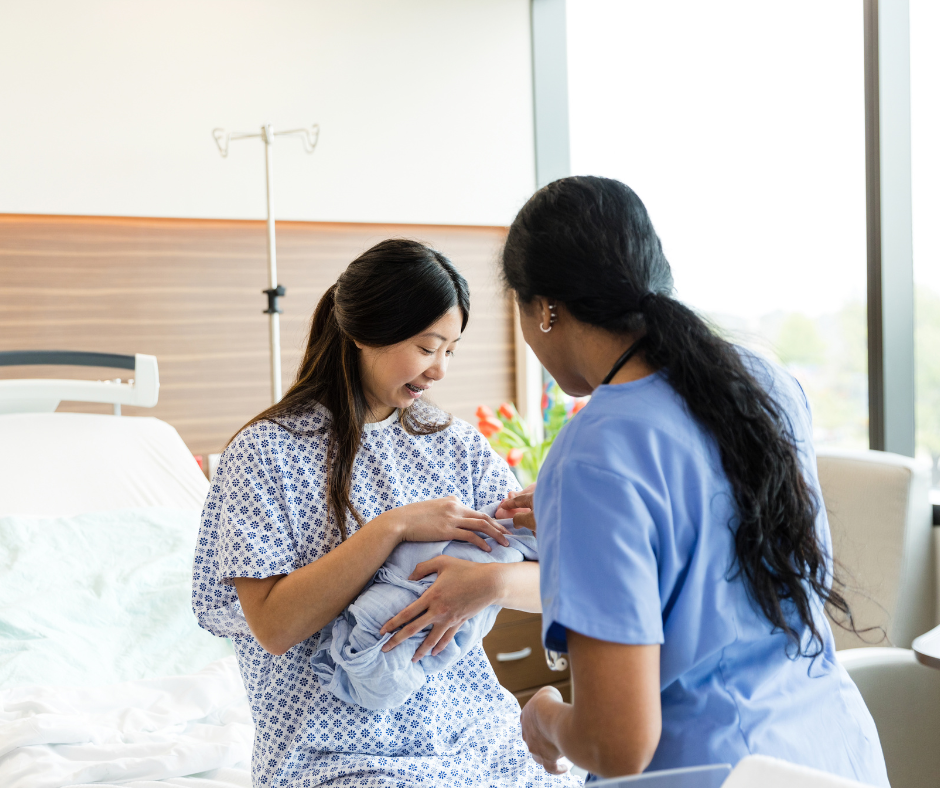 A person in a hospital gown holds a newborn infant while a healthcare professional in blue scrubs provides assistance.