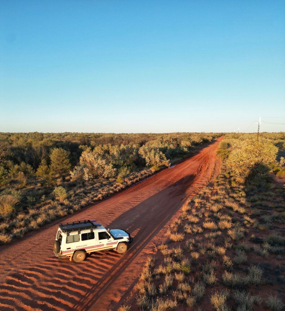A white off-road vehicle drives along a red dirt road through a sparse, dry Australian outback landscape.