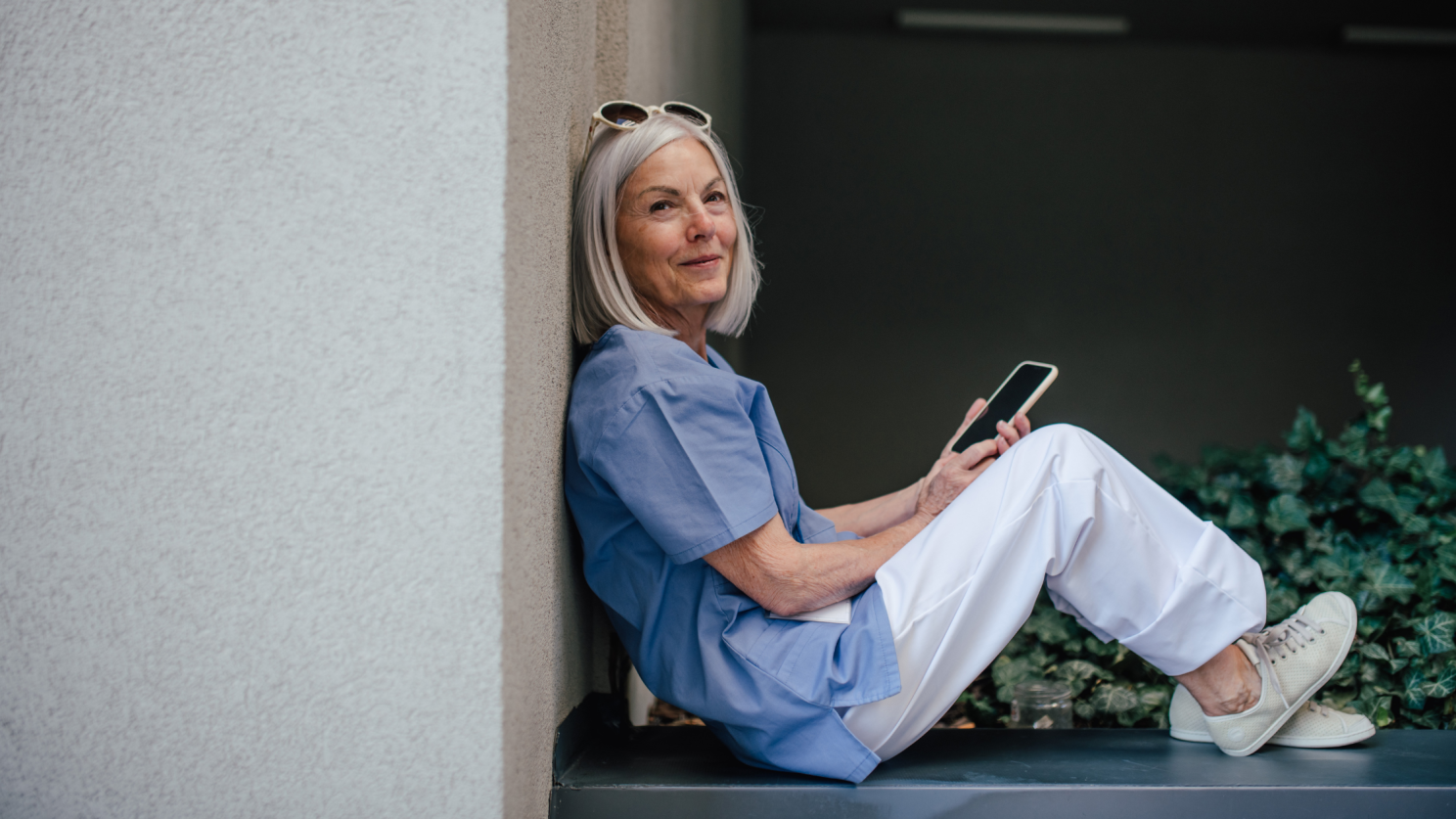 A person in blue scrubs and white pants sits leaning against a wall while looking at a smartphone.
