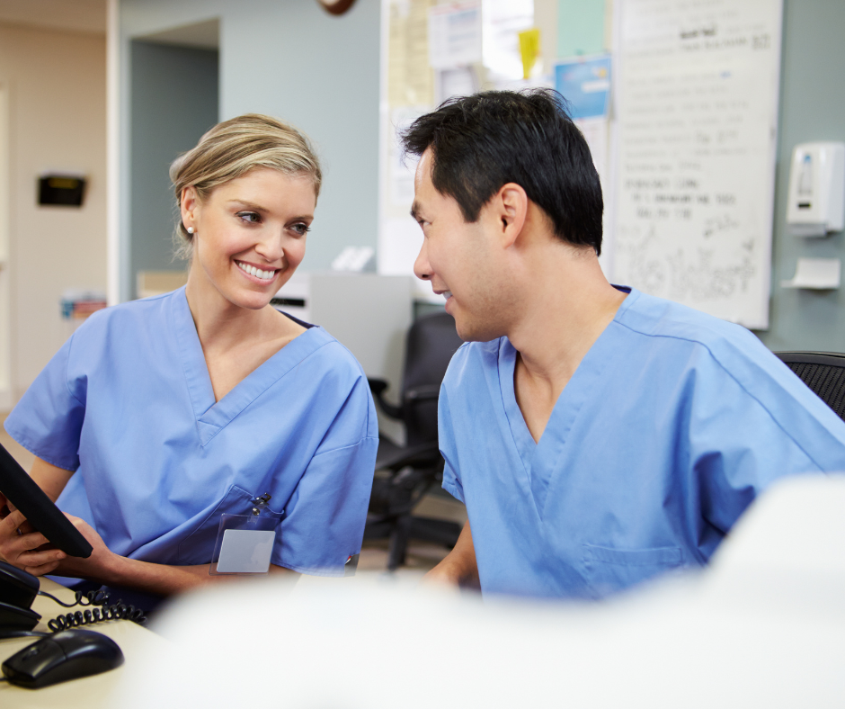 Two healthcare workers in blue scrubs look at a tablet together in a hospital office.