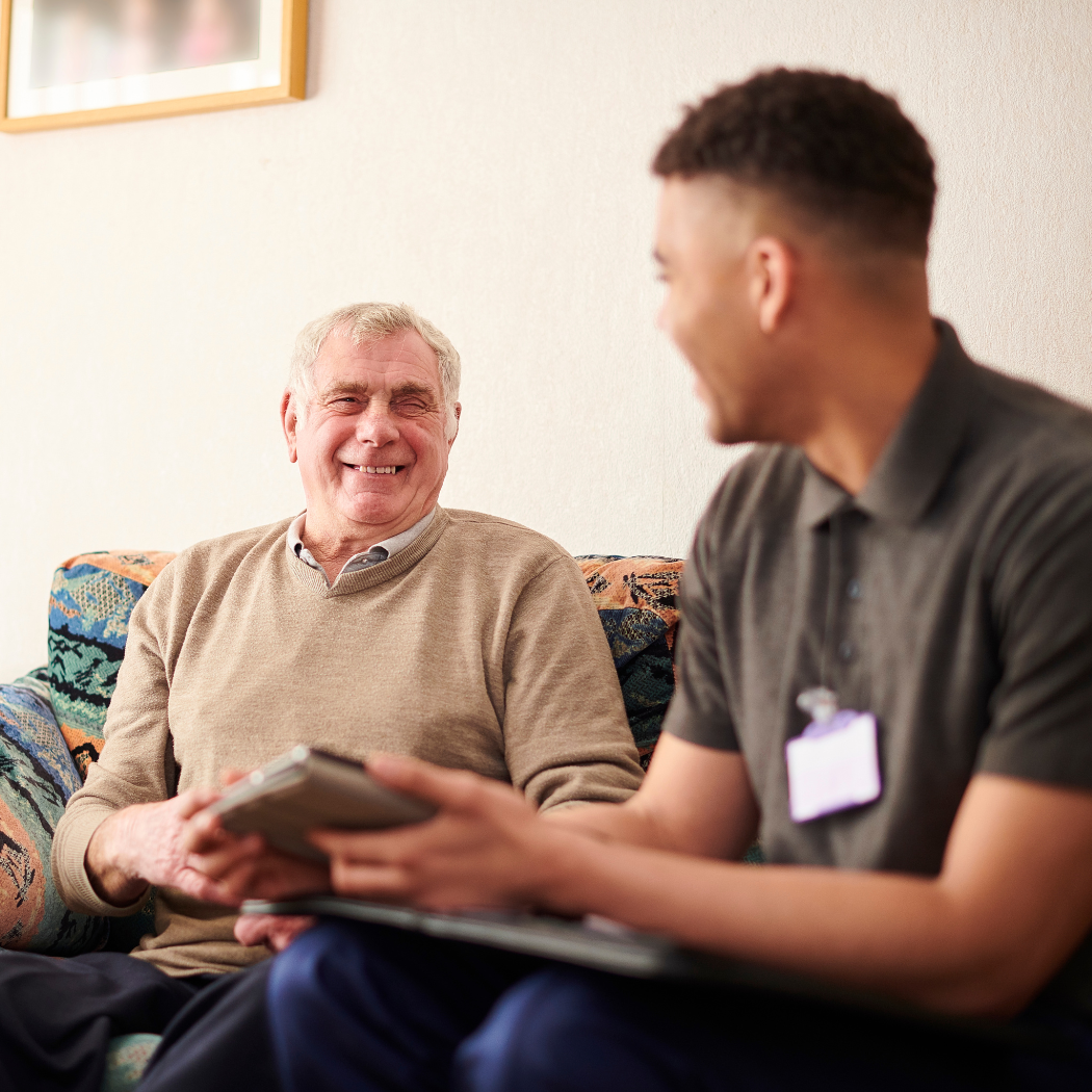 A person sitting on a couch shows a digital tablet to a smiling older individual.