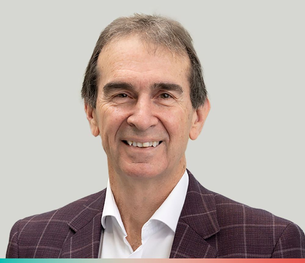 A person with short brown hair smiles while wearing a white collared shirt and a purple plaid blazer against a gray wall.