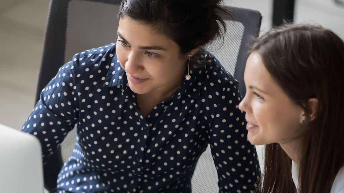 Two women in a bright office space collaborate while looking at a computer screen.