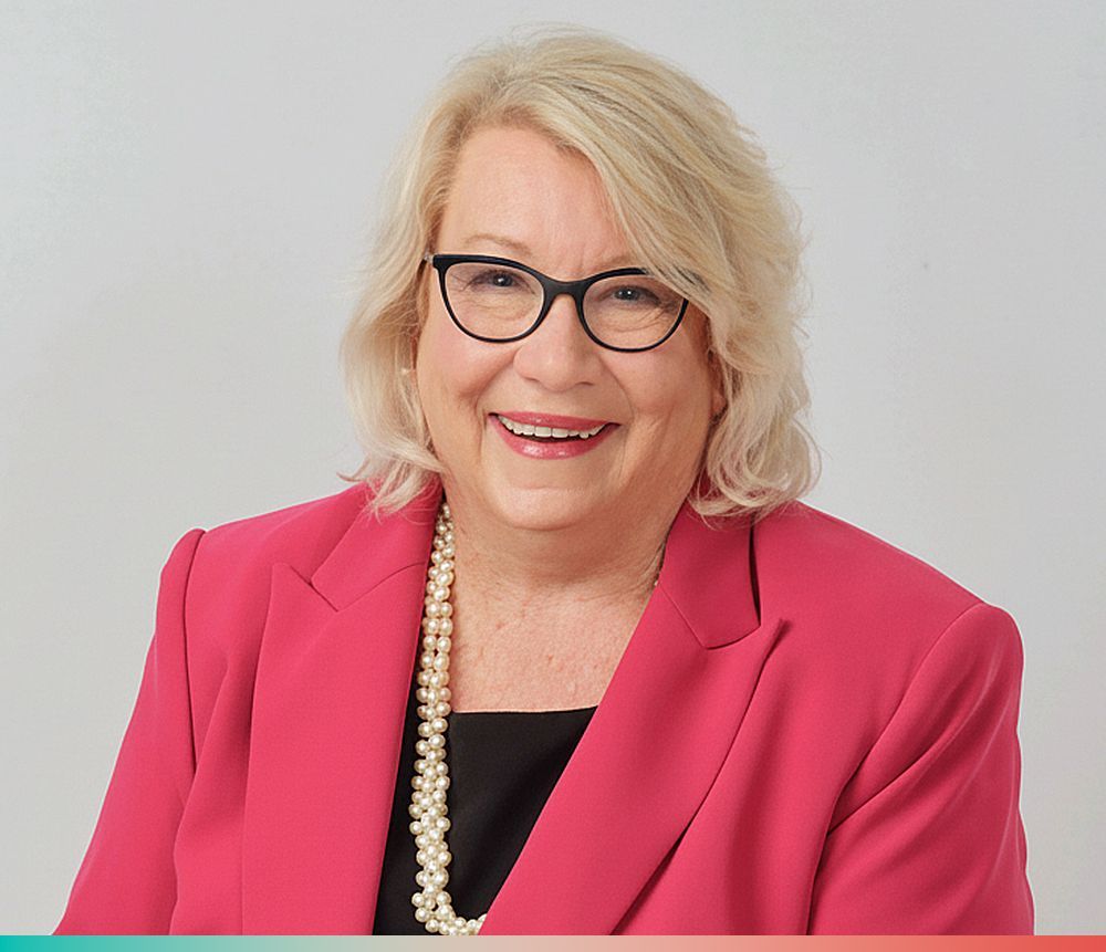 Smiling woman wearing black-rimmed glasses, a bright pink blazer, and a long pearl necklace against a light background.