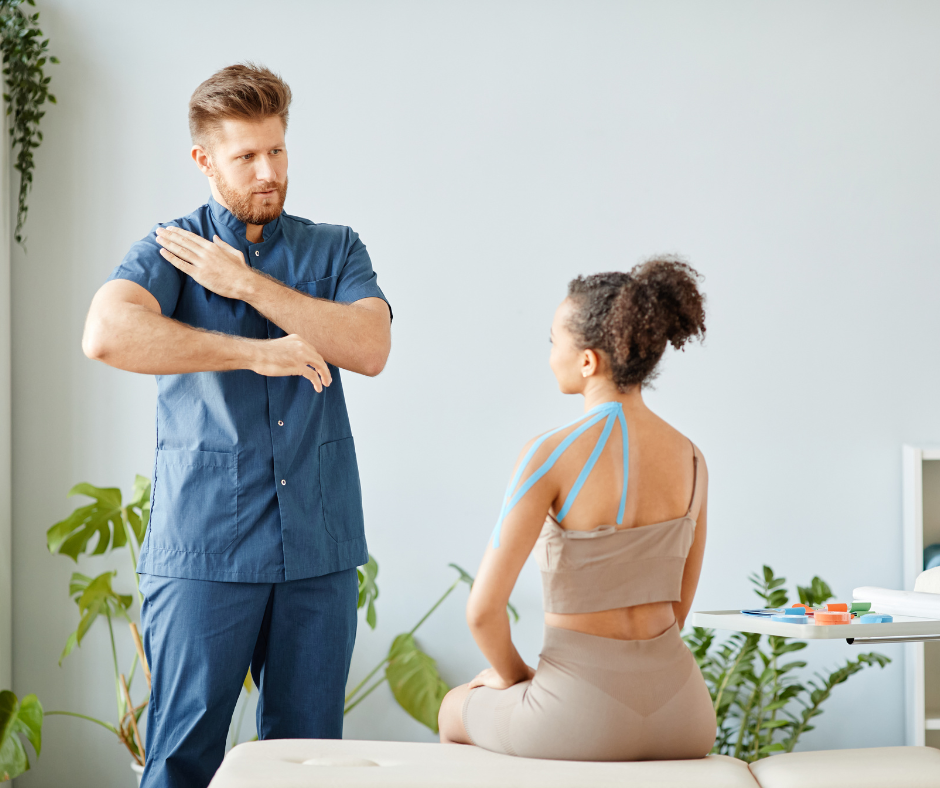 A physical therapist in blue scrubs demonstrates a shoulder exercise to a seated patient with kinesiology tape applied.
