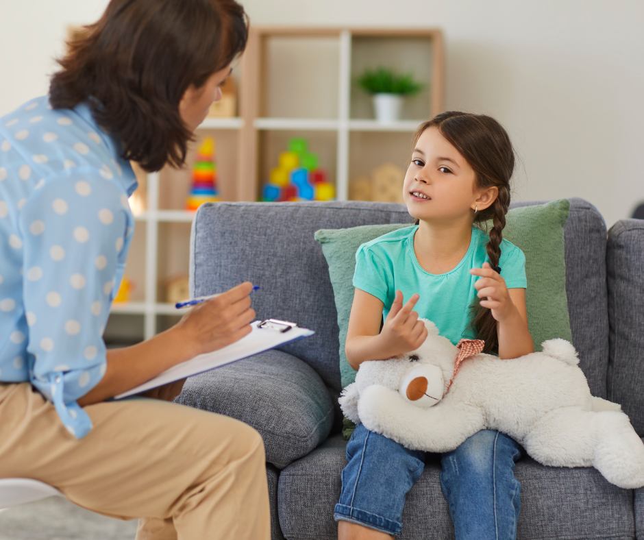 A therapist writing on a clipboard while a young girl with a plush toy speaks to them in a bright office.