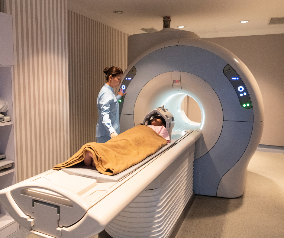 A patient lies under a brown blanket on an MRI machine table while a technician prepares the head scan.
