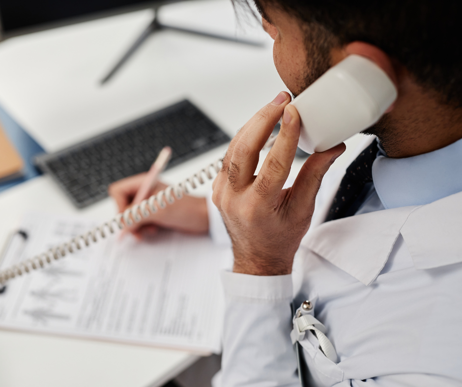 A healthcare professional in a white coat talks on a corded landline phone while writing notes on a medical form.