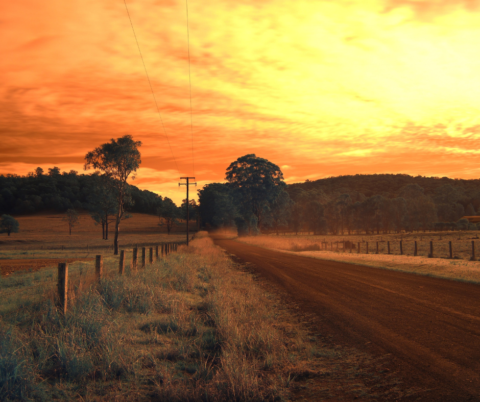A dirt road stretches toward a tree line under a vibrant, fiery orange sunset in a rural landscape with wooden fencing.