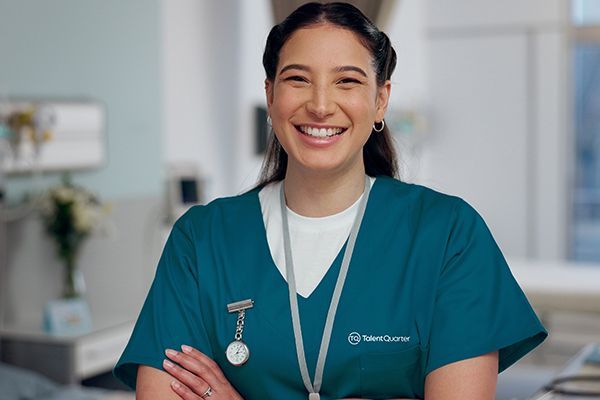 A smiling healthcare professional wearing teal scrubs and a lanyard stands with arms crossed in a clinical setting.