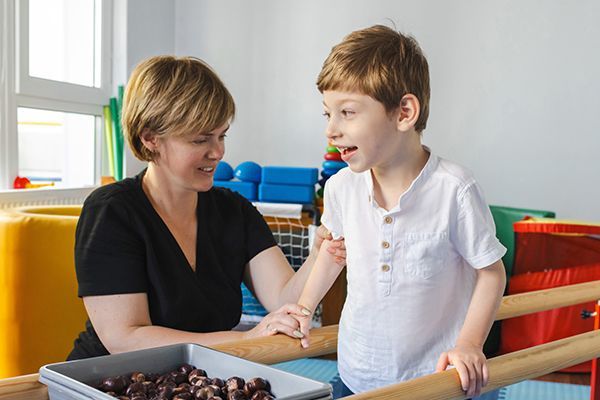 A person in a black shirt supports a child at parallel bars in a therapy room, with a tray of sensory objects nearby.