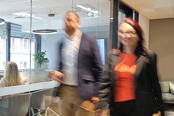 Two blurred people walking through an office past a glass-walled conference room with an indoor plant.