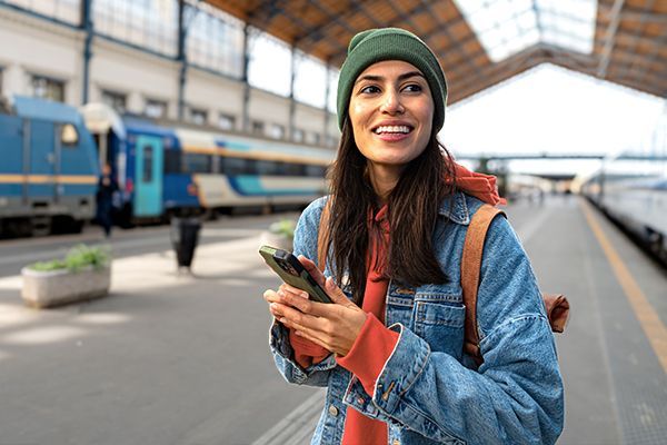 A smiling person in a beanie and denim jacket holds a smartphone while standing on a sunny train station platform.