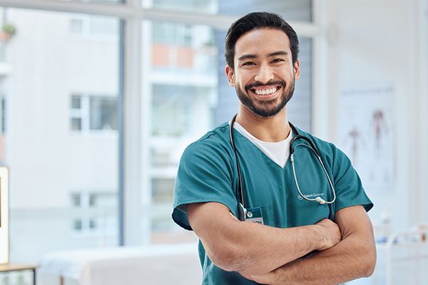 A smiling healthcare professional in green scrubs and a stethoscope standing with arms crossed in a clinic.