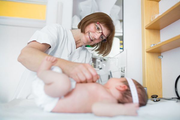 A medical professional in white scrubs smiles while adjusting a diaper on an infant lying on an exam table.