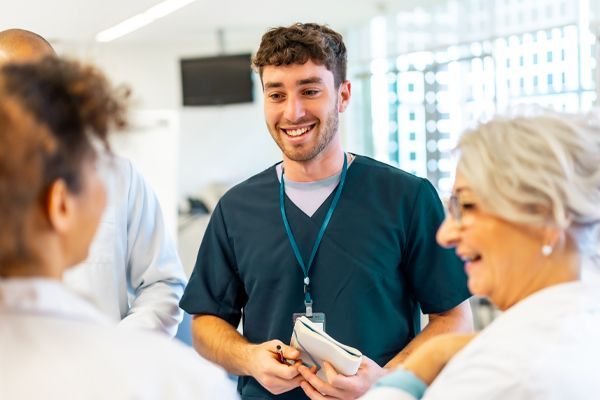 Three medical professionals in scrubs and lab coats talk and smile in a bright, modern clinical setting.