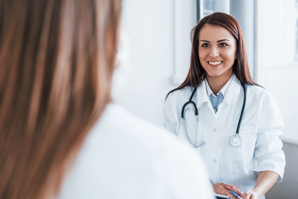 A smiling medical professional in a white coat with a stethoscope around their neck talks to a patient.