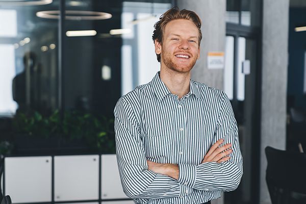 A professional standing with arms crossed, smiling, in a bright, modern office with glass partitions.