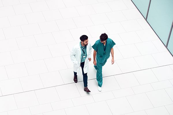 A doctor in a white coat and a colleague in green scrubs walk together across a white, tiled hallway.