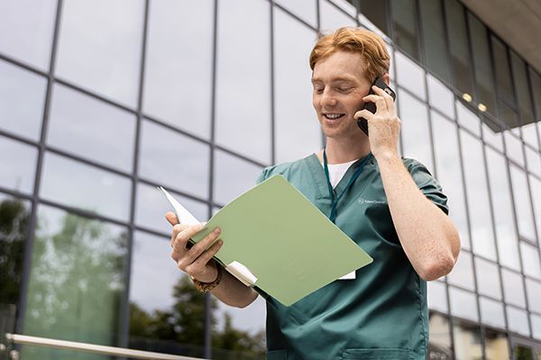 A person in green scrubs standing outside a modern building, reading a document while talking on a smartphone.