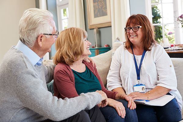 A care worker sits on a sofa, smiling and talking with an older couple in a bright, home-like setting.