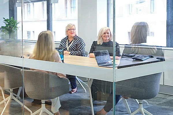 Four professionals sit at a light-colored wooden table in a glass-walled conference room during a meeting.