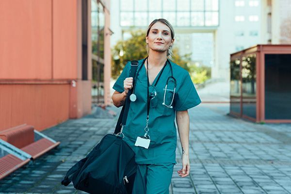 A healthcare professional in teal scrubs and a stethoscope walks outside a building, carrying a black bag.
