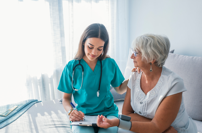 A healthcare worker in teal scrubs measures the blood pressure of a patient while documenting notes on a clipboard.